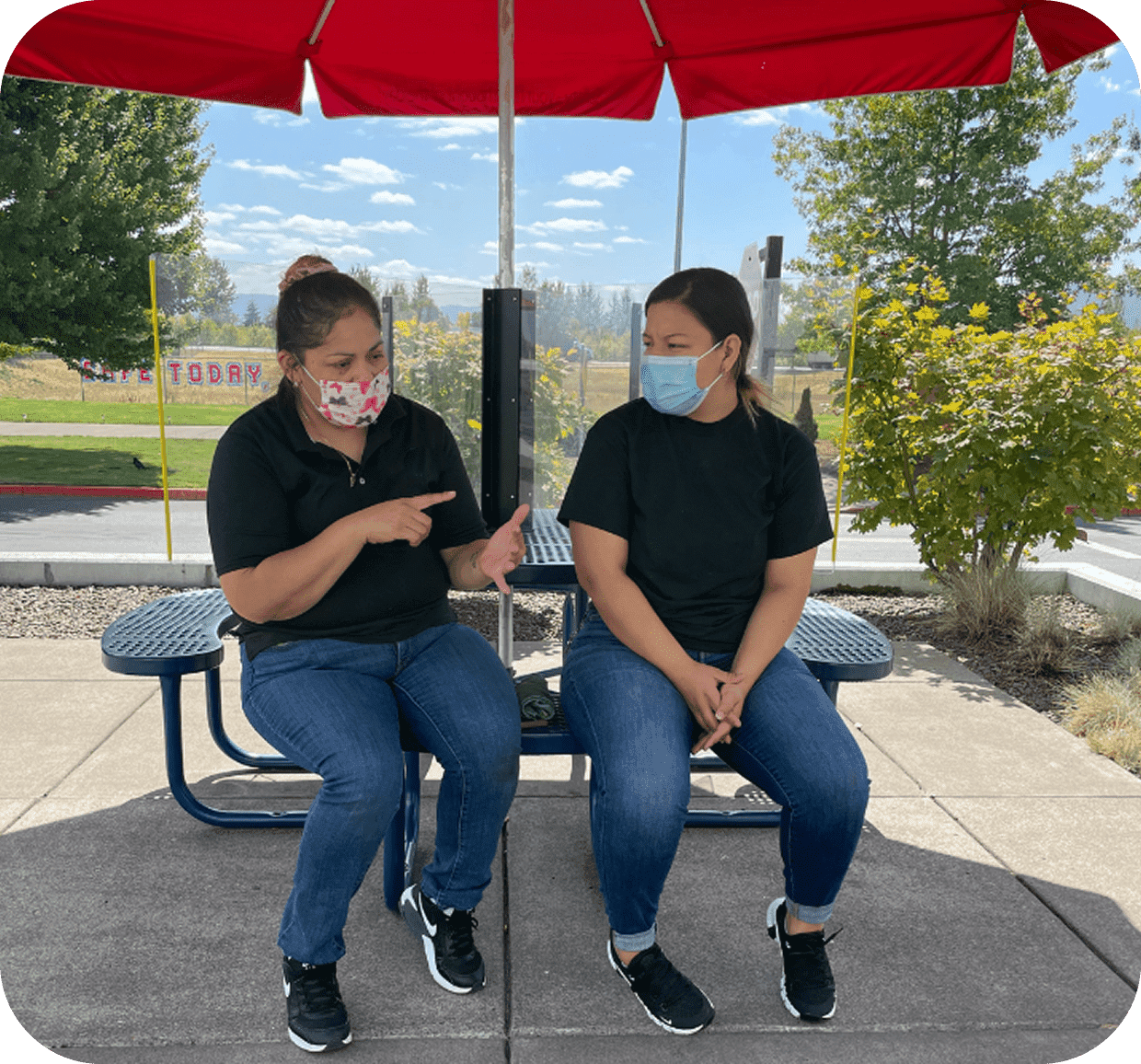 Two women wearing masks sitting under a red umbrella outdoors.